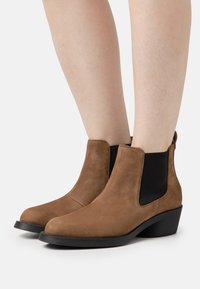 Botas de tobillo de ante marrón con paneles elásticos laterales negros, punta redonda y un tacón bajo tipo cuña, que presentan una textura suave y un diseño elegante.