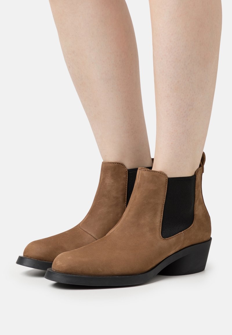 Botas de tobillo de ante marrón con paneles elásticos laterales negros, punta redonda y un tacón bajo tipo cuña, que presentan una textura suave y un diseño elegante.