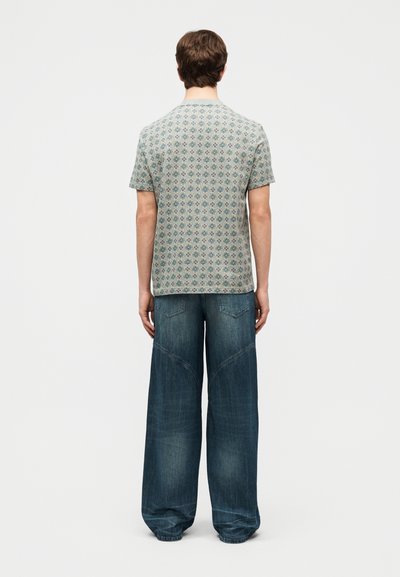 Άνδρας που στέκεται με την πλάτη γυρισμένη, φοράει κοντομάνικο πουκάμισο με σχέδια και φαρδιά μπλε τζιν παντελόνια, πάνω σε απλό φόντο.