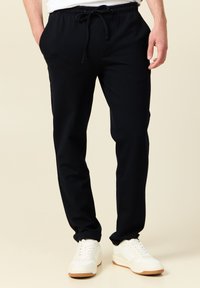 Pantalones de chándal negros hechos de una mezcla de algodón, con cintura elástica y cordón ajustable, y bolsillos laterales. Textura suave con piernas ajustadas.