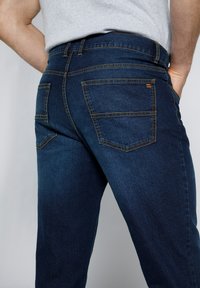 Jeans de denim en azul oscuro con una textura suave. Presentan cinco bolsillos, costuras naranjas en contraste y un diseño ajustado que se estrecha en el tobillo.