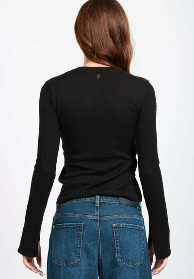 Donna con lunghi capelli castani che indossa una maglietta aderente nera a maniche lunghe e jeans blu a vita alta, in piedi con la schiena rivolta verso la fotocamera.