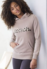 Beige Kapuzenpulli mit dem Logo "Bench." in Schwarz und Weiß, mit einer Kängurutasche und gerippten Bündchen. Kombiniert mit dunklen Leggings.