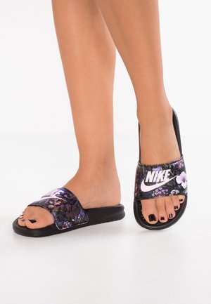 Personne portant des claquettes Nike noires avec des motifs floraux violets et des ongles d’orteils peints en noir sur fond blanc.