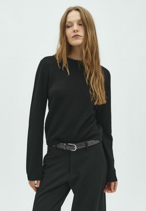 Jeune femme aux longs cheveux blonds porte un pull noir, un pantalon noir et une ceinture noire, debout devant un fond clair uni.