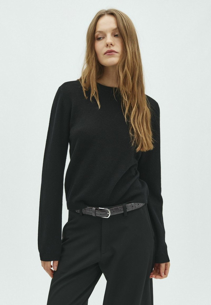 Jeune femme aux longs cheveux blonds porte un pull noir, un pantalon noir et une ceinture noire, debout devant un fond clair uni.