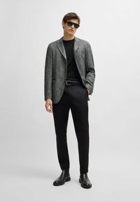Blazer grigio a spina di pesce con accenti neri, abbinato a un top nero aderente e pantaloni neri su misura. Gli stivaletti neri alla caviglia completano il look.