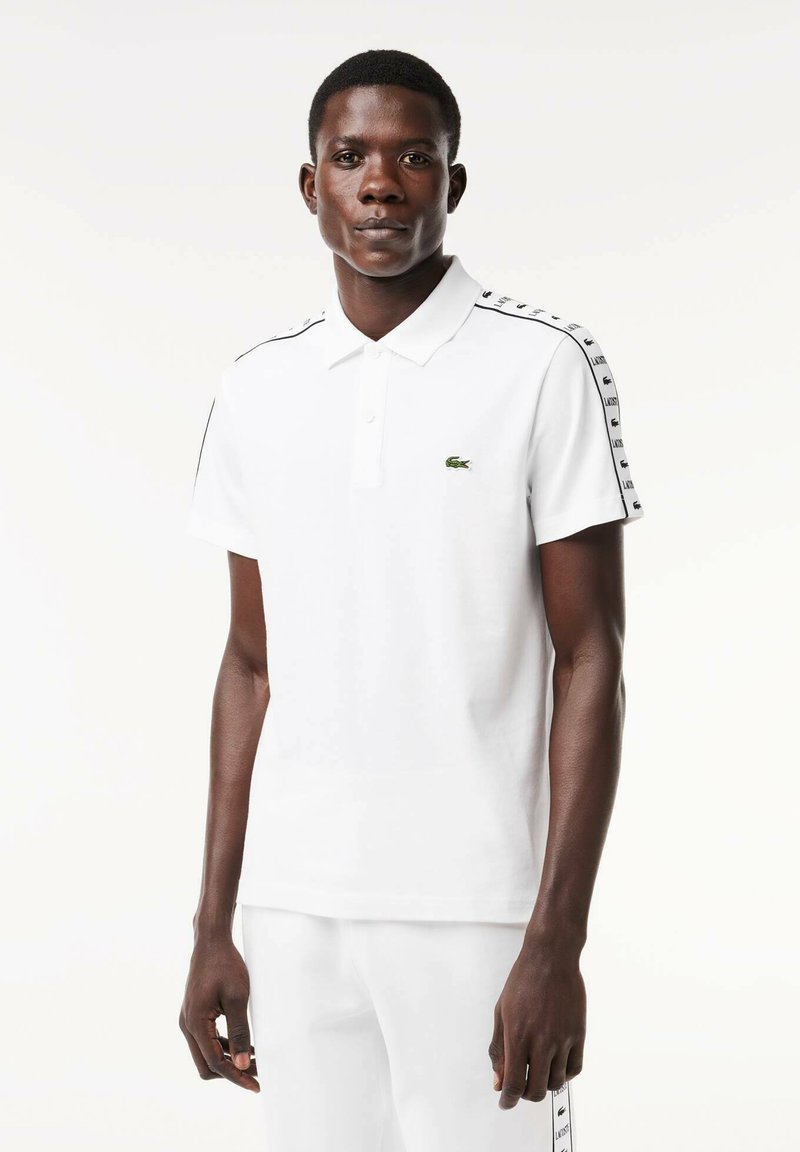 Polo shirt blanc à manches courtes, arborant un logo vert sur la poitrine et des accents noirs le long des épaules.