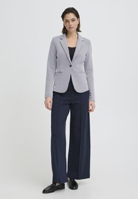Veste blazer gris clair sur mesure avec revers crantés, fermeture par un bouton unique et poches avant, associée à un pantalon large bleu marine et des mocassins noirs.