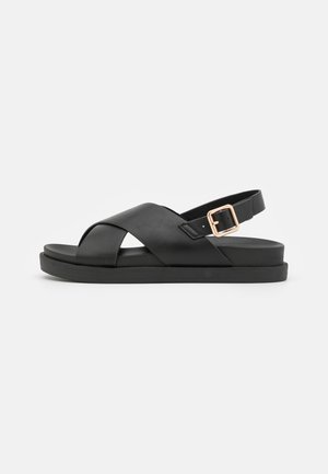 Sort flad sandal med korslagte læderremme og justerbar ankelrem med guldlås på en ensartet lys baggrund.