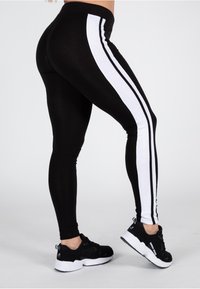 Leggings neri con strisce bianche laterali, realizzati in materiale elasticizzato. Presentano un alto punto vita e una superficie liscia. Abbinati a scarpe sportive nere.