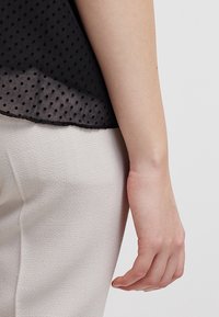 Zwarte blouse met stippenpatroon en een doorschijnende zoom, gecombineerd met beige broek met textuur, met een hand rustend op de dij.