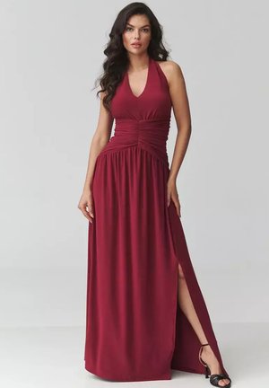 Kobieta o długich ciemnych włosach, ubrana w bezrękawnikową bordową maxi sukienkę z kołnierzem halter i rozcięciem po boku oraz czarne sandały na wysokim obcasie.