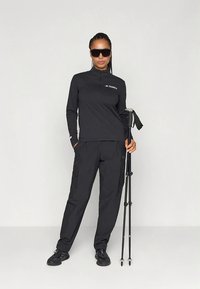 Must musta välisjak, millel on pool lukku, ühendatud mustade cargo-pükste ja matkakeppidega. Modell kannab päikeseprille ja musti tossusid.