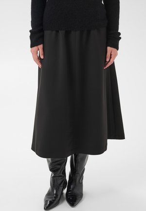 Femme portant une jupe noire au genou, un pull en maille noir et des bottes noires brillantes à bout pointu, debout sur un fond blanc.