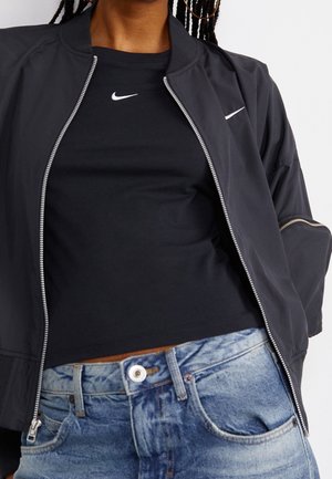 Person trägt eine schwarze Nike Jacke mit Reißverschluss über einem schwarzen Nike T-Shirt, kombiniert mit hoch taillierten hellblauen Jeans.