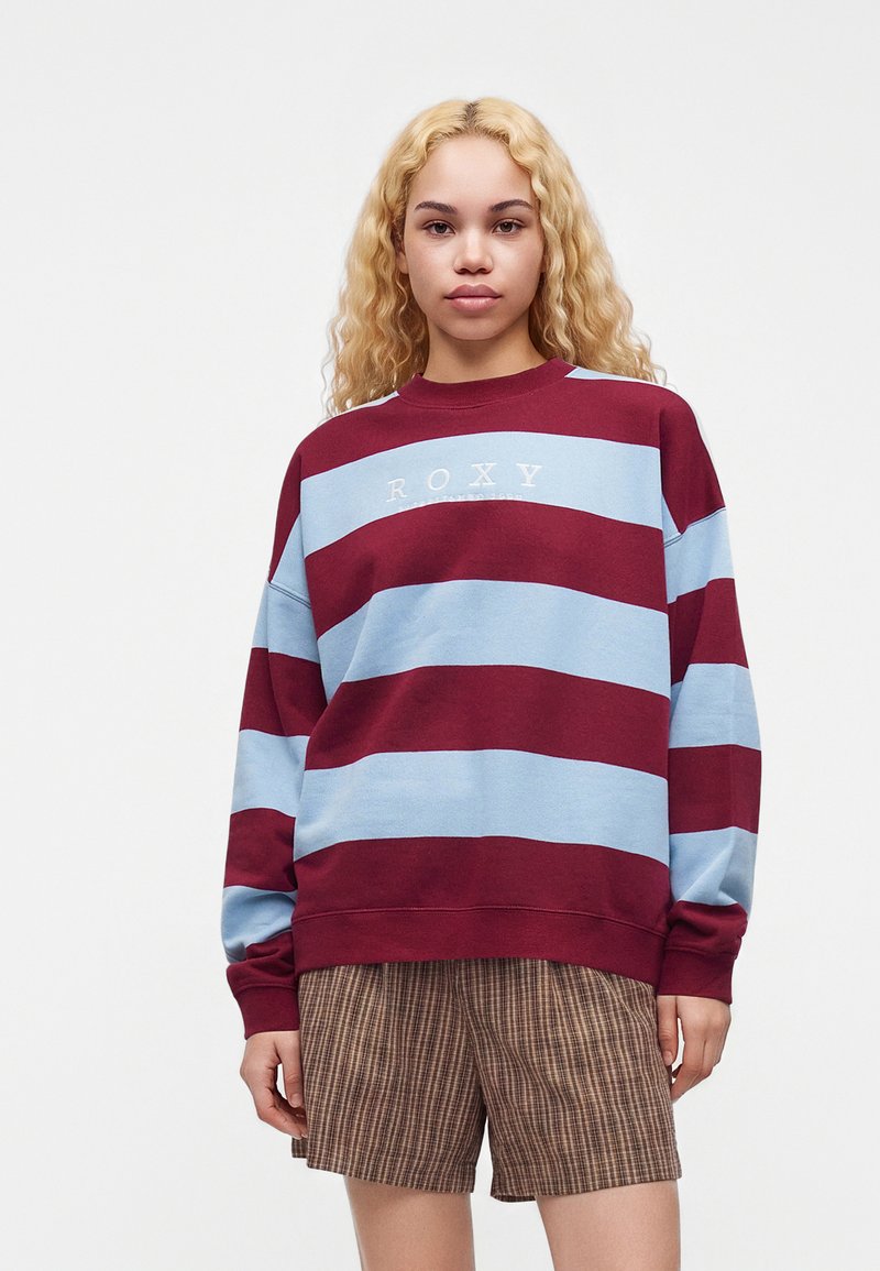 Jeune femme aux longs cheveux blonds bouclés portant un pull rayé bordeaux et bleu clair et un short marron à carreaux sur fond blanc.