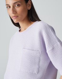Felpa texturizzata di un leggero colore lavanda con motivo a coste, caratterizzata da una tasca frontale e maniche ampie. Materiale in cotone morbido evidente.