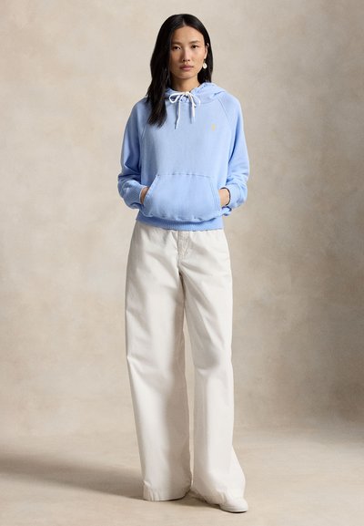 Jeune femme portant un sweat à capuche bleu clair avec poche frontale et un pantalon large crème, debout devant un fond beige neutre.