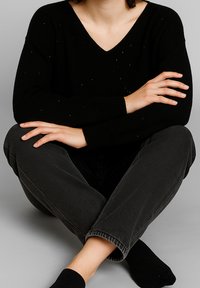 Maglione nero a collo a V con piccoli accenti decorativi in argento, abbinato a jeans grigio scuro, in posa seduta, con le braccia incrociate e piedi nudi.