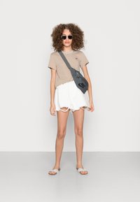 T-shirt riscado em castanho e bege, calções brancos desgastados, mala cinza de ombro com logotipo preto, e chinelos cinzas com um detalhe de laço.