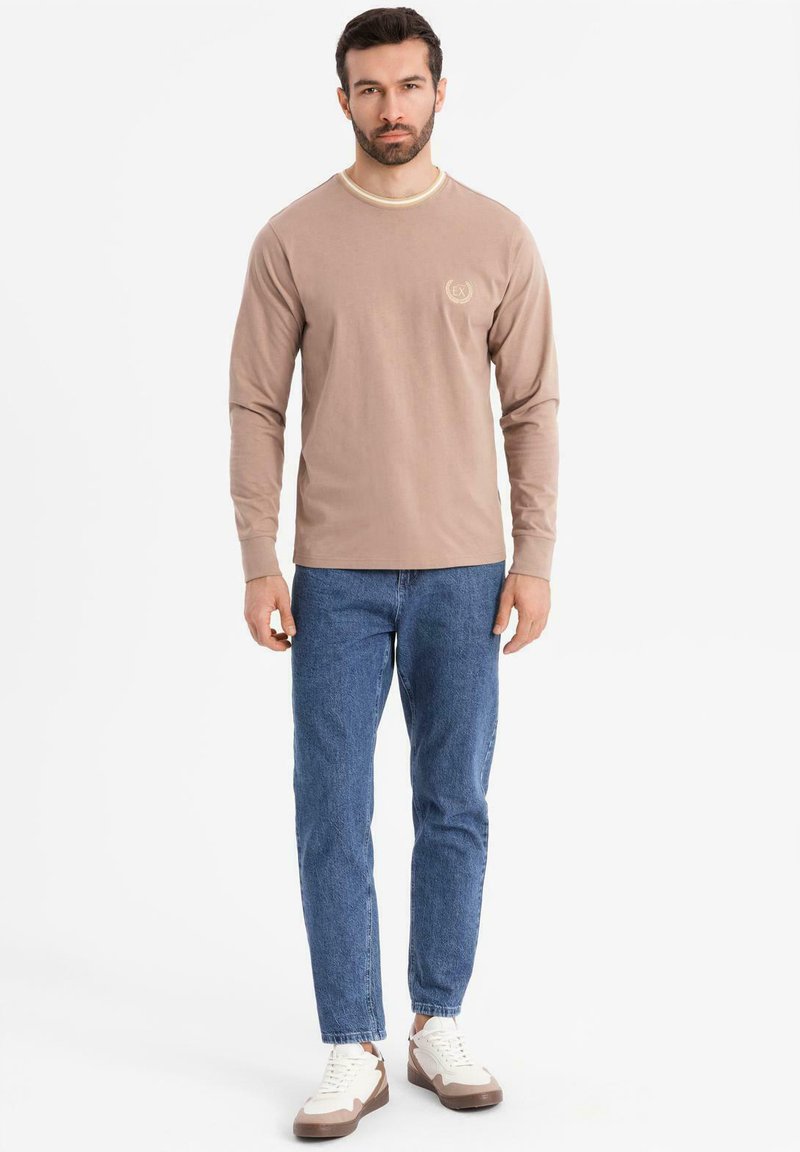 Beige Langarmhemd mit rundem Ausschnitt und hellfarbigem Besatz, versehen mit einem kleinen Logo. Kombiniert mit blauen Jeans und Sneakern.