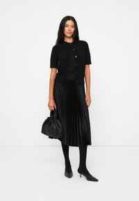 Cardigan nero a maniche corte con bottoni, abbinato a una gonna nera plissettata, collant neri, borsa nera e tacchi a punta neri.