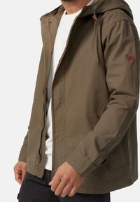 Chaqueta verde oliva con capucha, con cierre de cremallera, puños elásticos y un parche de cuero marrón con el logo en el hombro izquierdo.