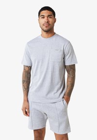 Camiseta gris con cuello redondo y un bolsillo en el pecho. La tela parece suave, con mangas cortas y un corte relajado, combinada con pantalones cortos grises.