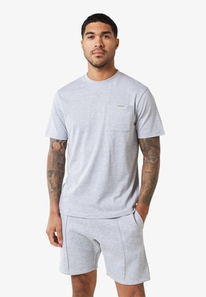 T-shirt grigia con scollatura tonda e taschino sul petto. Il tessuto sembra morbido, con maniche corte e una vestibilità comoda, abbinato a pantaloni corti grigi.