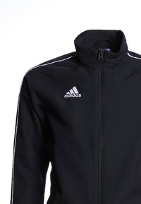 Sort atletisk jakke med høj krave, hvide striber langs skuldrene og et sølvfarvet adidas-logo på venstre bryst. Lynlås foran.