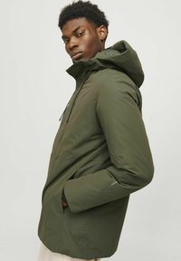 Chaqueta con capucha de color verde oliva, con una textura suave, puños elásticos y bolsillos frontales; incluye cordones ajustables y un diseño minimalista.
