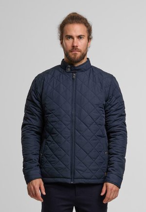 Laivaston sininen tikattu takki, jossa on pystysuora kaulus, vinopistot, edessä vetoketju ja sivutaskut. Kangas on sileä ja mattapintainen.