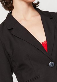 Svart blazer i strukturerad tyg, med spetsig krage, enkelknäppning och subtila fickdetaljer.