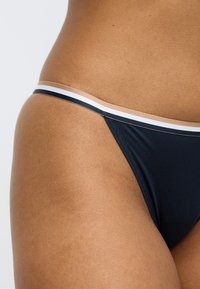 Merensinisen bikinihousut, joissa on sileä pinta ja beige-valkoraidallinen vyötärönauha. Muotoilu on pelkistetty ja alaosa on matalavyötäröinen.