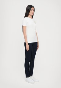 T-shirt branca de manga curta com decote redondo e um logótipo no peito, combinada com jeans azul escuro e ténis brancos.