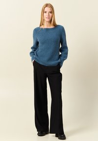 Pull bleu tricoté avec une texture côtelée, coupe décontractée et encolure bateau, associé à un pantalon large noir et des bottes noires épaisses.