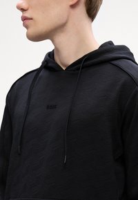 Μαύρη φούτερ κουκούλα με υφή γεωμετρικού σχεδίου, κορδόνια και ένα μικρό ανάγλυφο λογότυπο μπροστά.