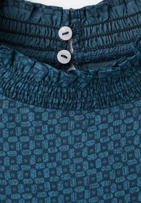 Teflă în tonuri închise de teal cu un model texturat în rețea, având un guler volan și două butoane albe la spate pentru închidere.