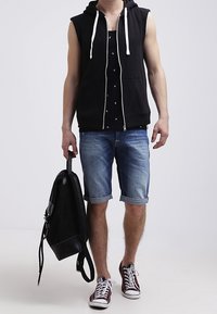 Sudadera sin mangas negra, camiseta gráfica negra, pantalones cortos de mezclilla y zapatillas burdeos. Mochila negra colgada sobre el hombro izquierdo.