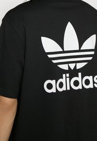 Černé bavlněné tričko s velkým bílým logem Adidas a třemi vodorovnými pruhy nad ním na zadní straně. Jednoduchý, klasický design.