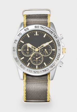 Chronograph watch with a black dial, silver bezel, yellow accents, and a textured gray fabric strap. Features tachymeter and multiple subdials.