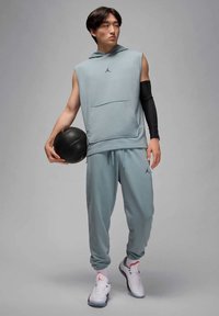 Mlad moški v brezrokavnem kapucarju in trenirki, ki drži črno košarkarsko žogo, nosi bele superge in črno rokavico na roki, stoji na sivi podlagi.