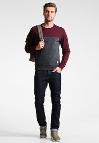 Sudadera de manga larga en color burdeos y gris con cuello redondo, combinada con jeans oscuros y zapatillas grises. Llevando una mochila marrón claro.