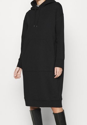 Osoba nosząca długą czarną sukienkę z kapturem typu hoodie, kieszenią kangurzą, ściągaczem przy kapturze i czarne kozaki za kolano na jednolitym jasnym tle.