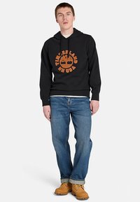 Czarna bluza z kapturem z pomarańczowym logo Timberland, w zestawieniu z niebieskimi dżinsami i brązowymi butami. Bluza wyposażona w przednią kieszeń i regulowane sznurki przy kapturze.