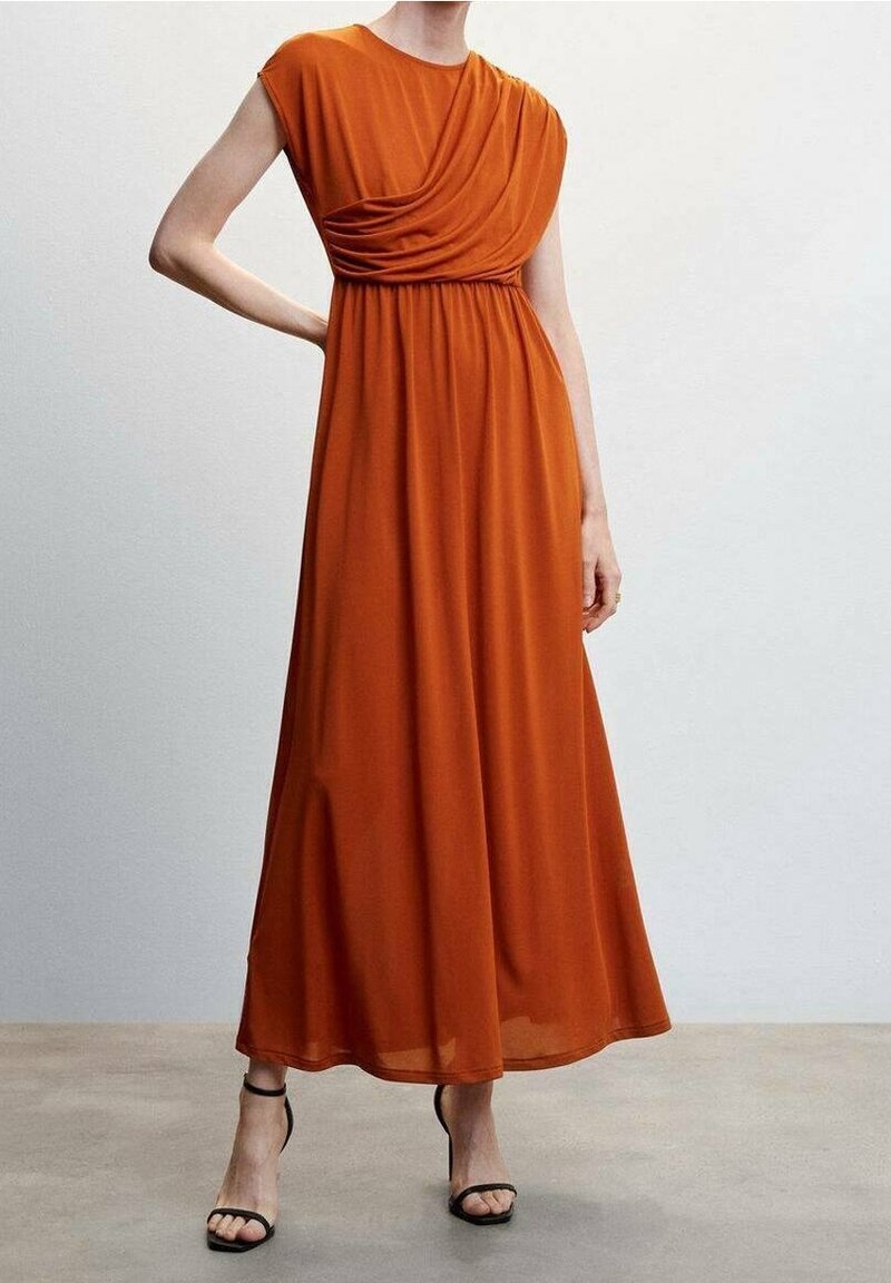 Femme portant une robe maxi sans manches orange rouille avec un corsage drapé et des talons ouverts noirs, debout contre un mur clair uni.