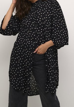 Femme portant une tunique noire à pois à manches longues par-dessus un jean noir, une main touchant ses cheveux et l'autre dans la poche du jean.