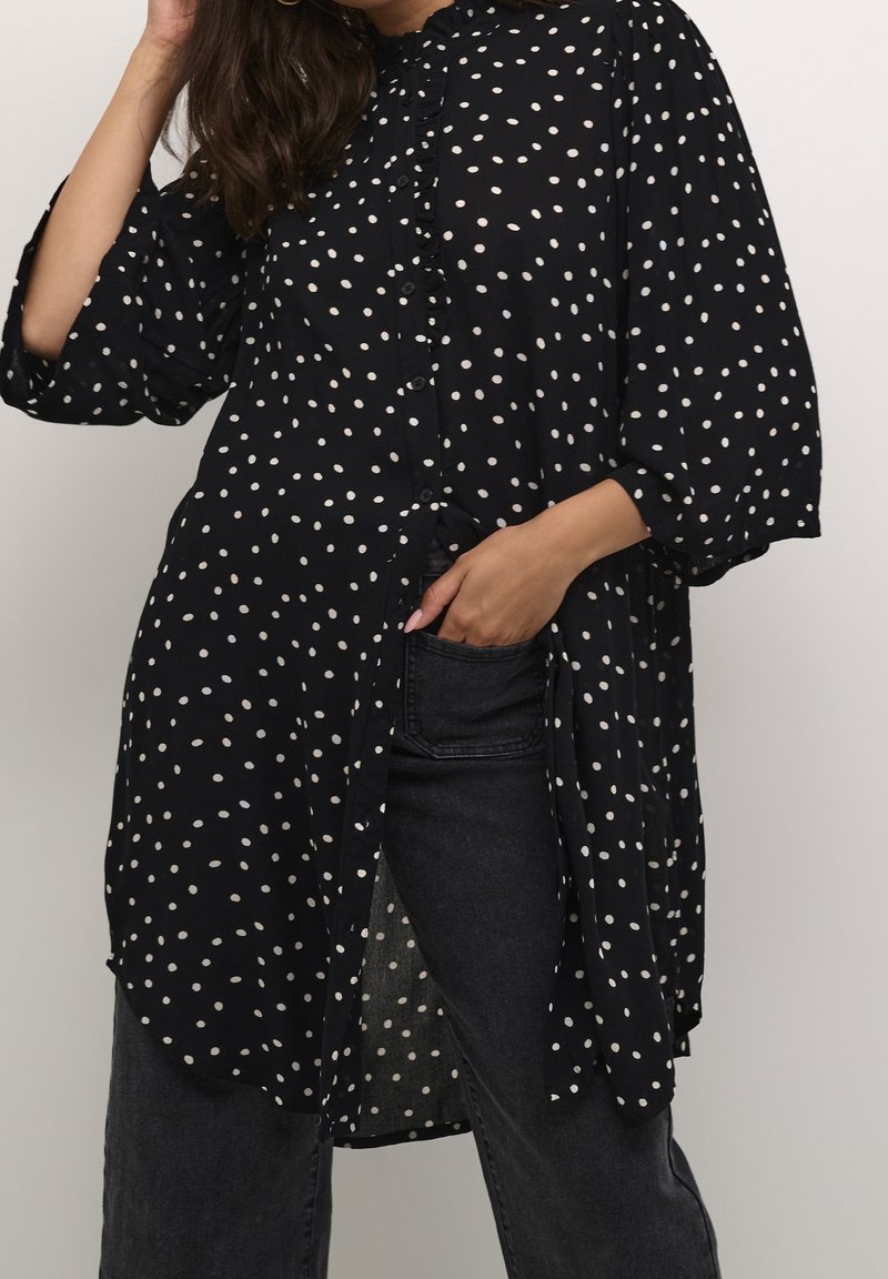 Femme portant une tunique noire à pois à manches longues par-dessus un jean noir, une main touchant ses cheveux et l'autre dans la poche du jean.