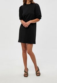 Robe noire à manches courtes avec une coupe décontractée, ornée de manches bouffantes et d'un col arrondi, associée à des sandales à talons vert olive.